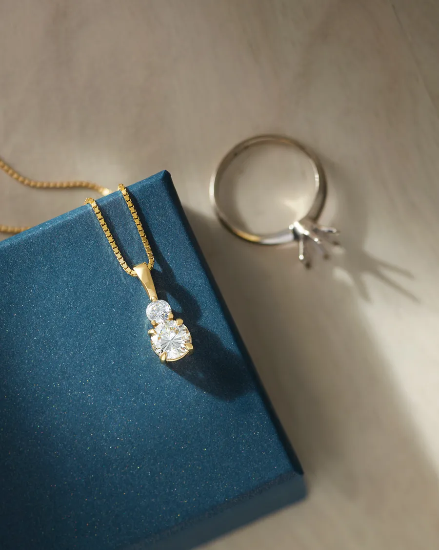 Gold necklace with two sparkling diamonds on a blue box, alongside a silver ring.