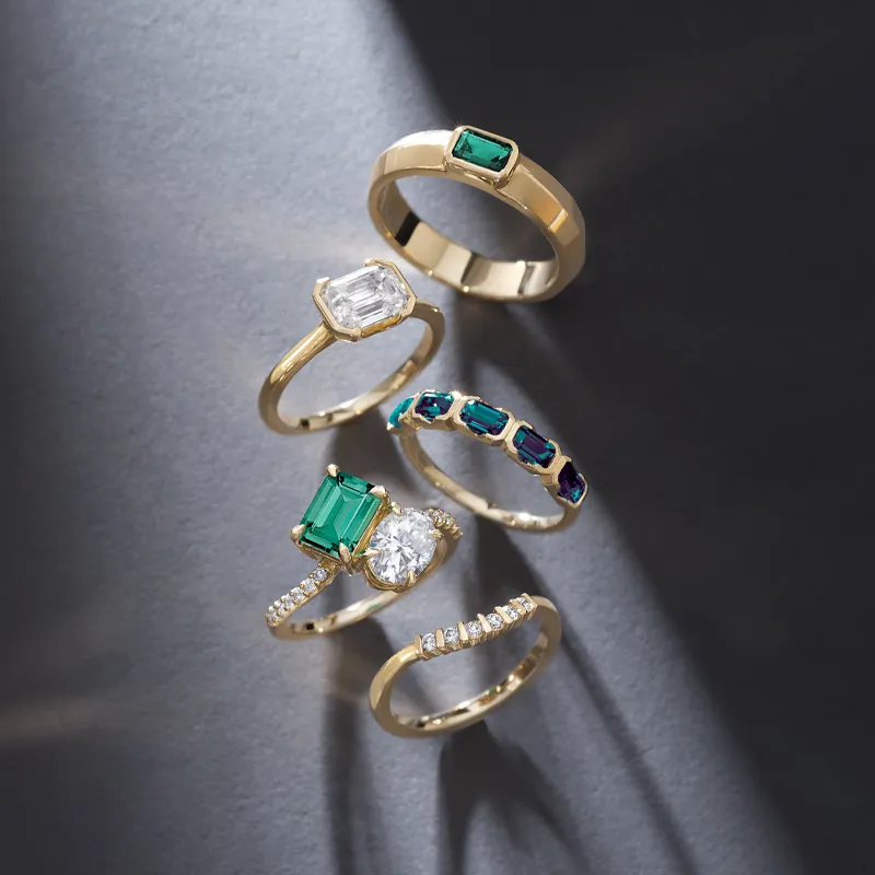 Five elegant gold rings featuring various gemstones, including emeralds and diamonds, displayed on a dark background.
