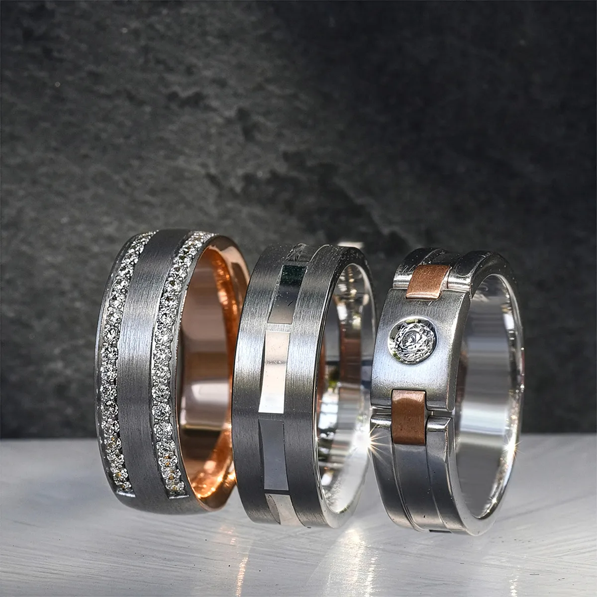 Three men's wedding bands with unique designs, featuring diamonds and mixed metals, displayed on a textured surface.