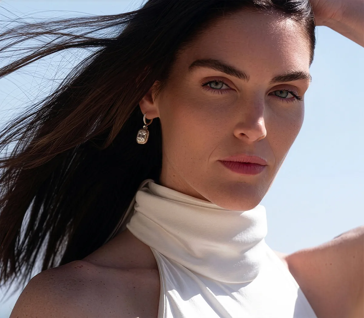 A woman with long hair wearing a white top and earrings, looking at the camera.