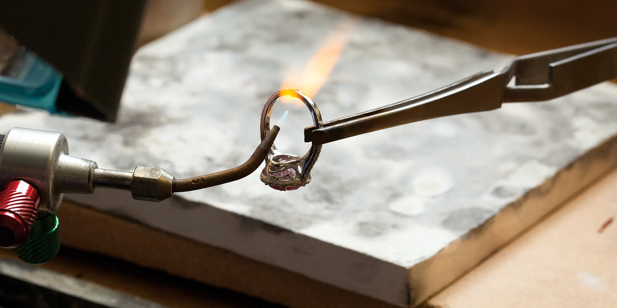 A jeweler uses a torch to work on a ring held by tweezers.