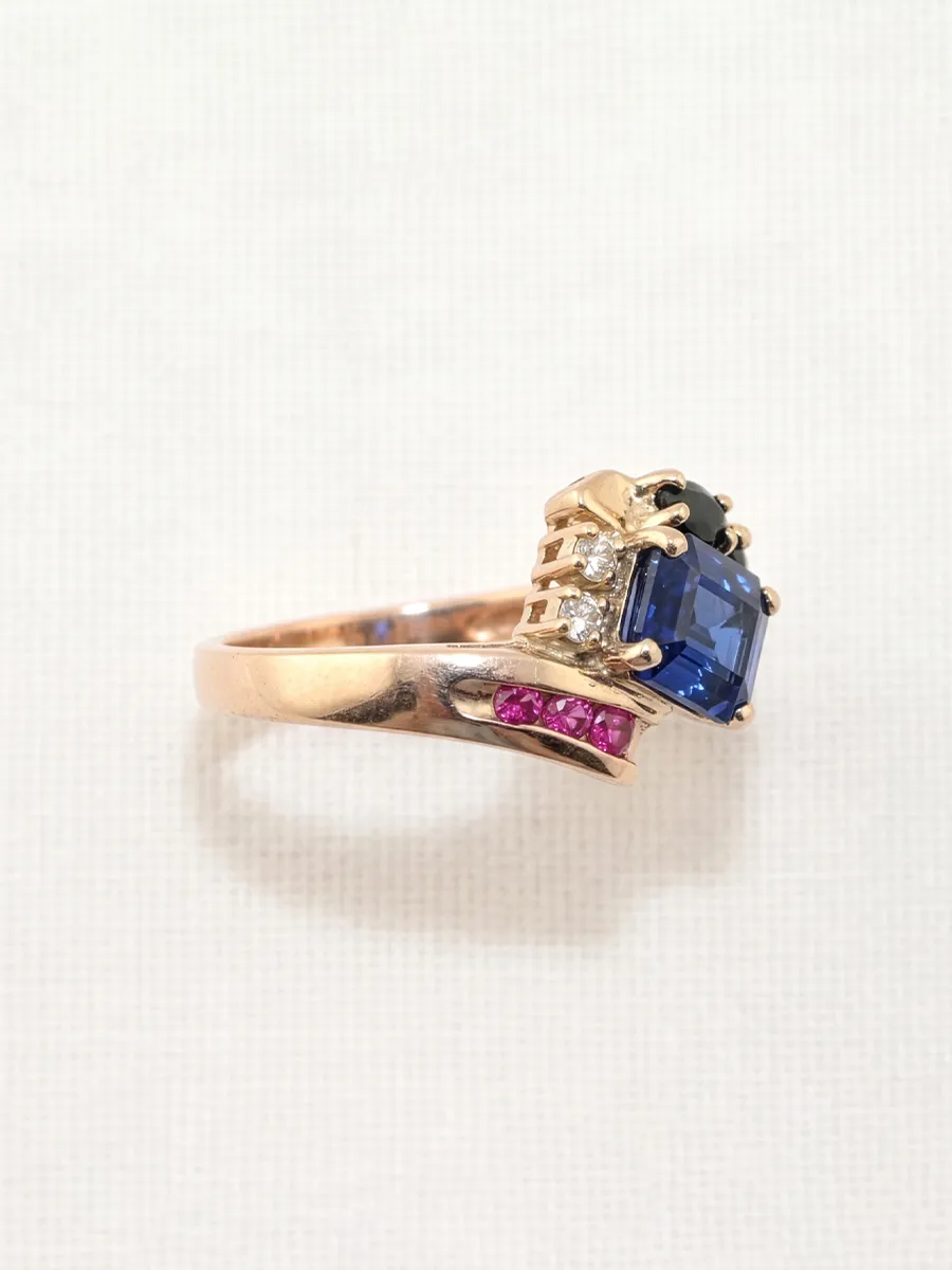 Gold ring with blue, pink, and clear gemstones on a light background.