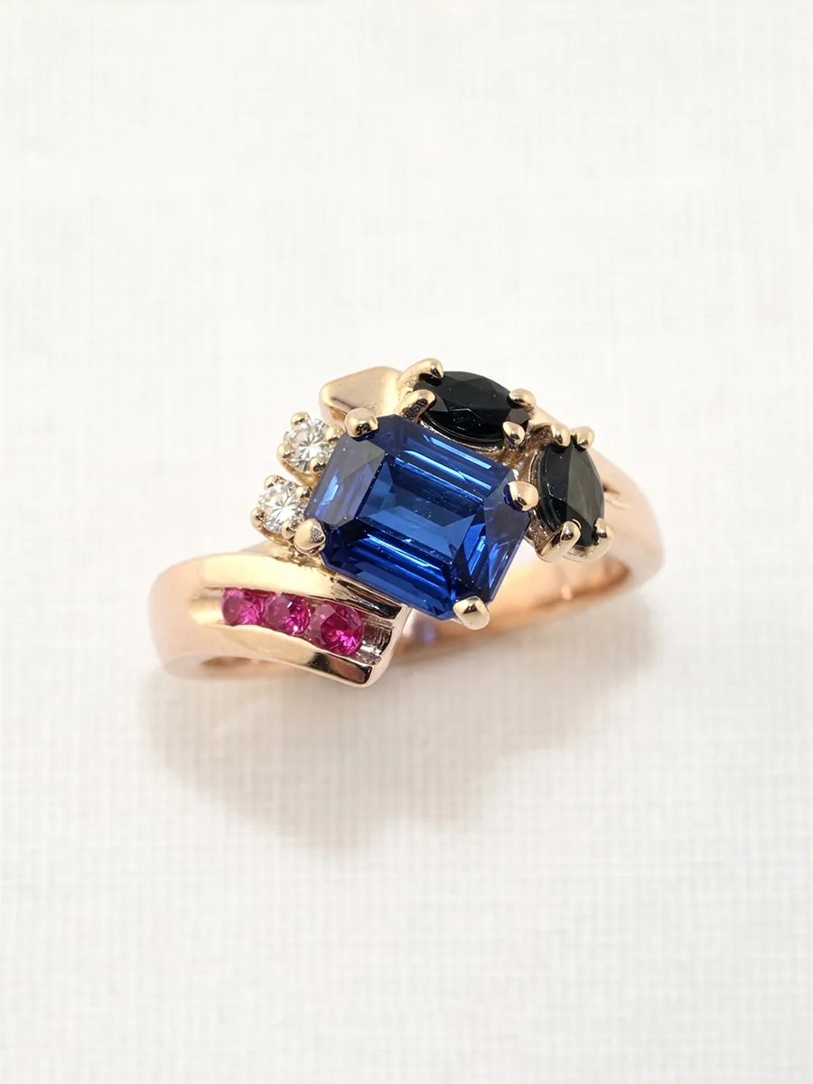 Gold ring with a blue gemstone, black stones, and small pink and white gems on a light background.
