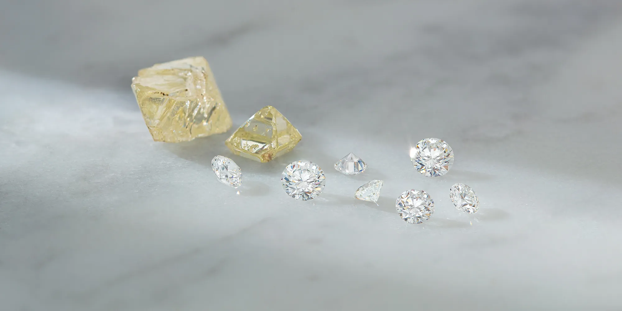 Uncut and cut diamonds displayed on a marble surface.
