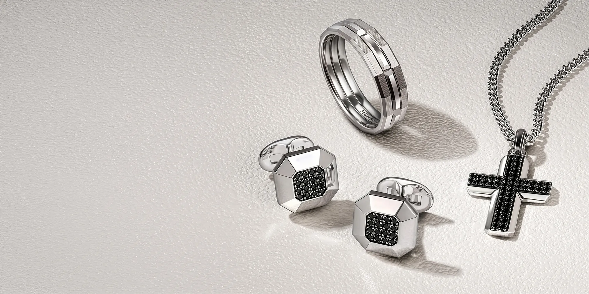 Silver ring, cufflinks, and cross pendant with black accents on a textured surface.
