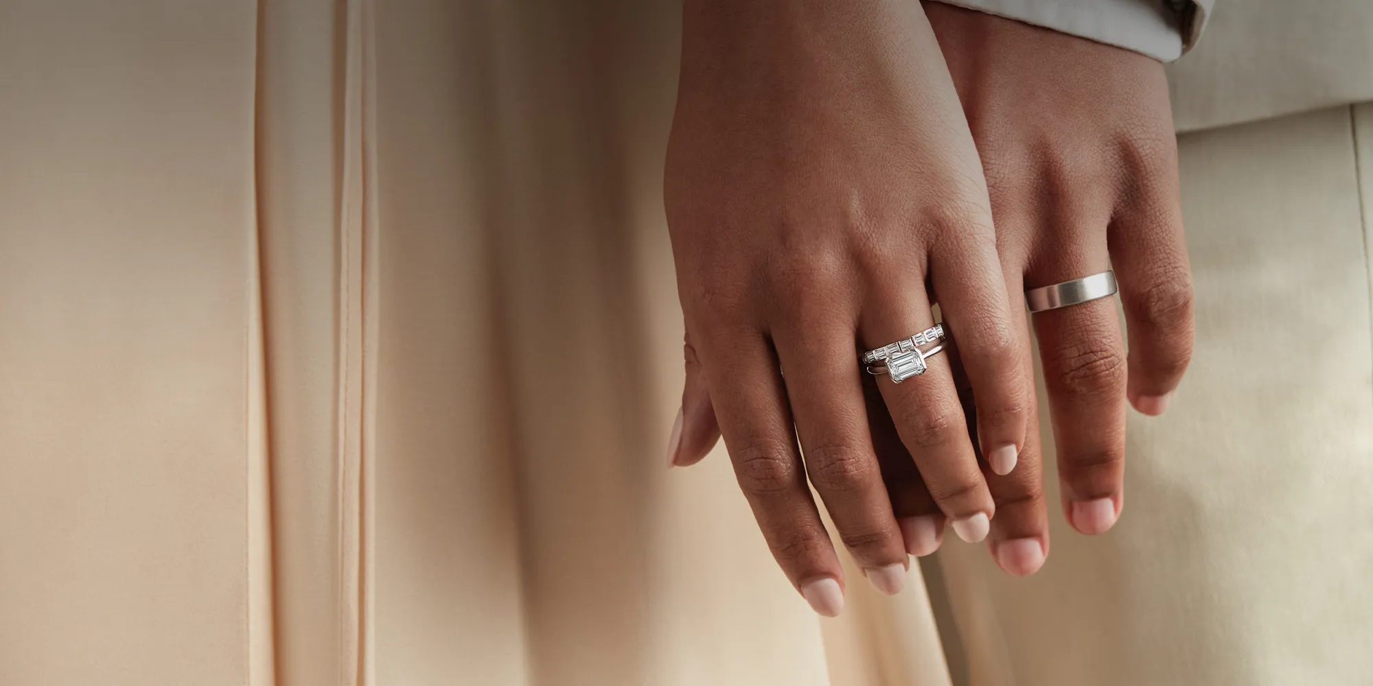 Hands with engagement rings, one with a diamond band, against a beige background.