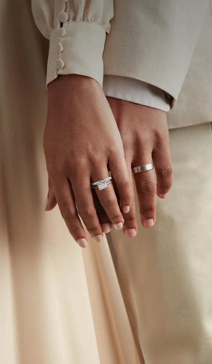 Hands with engagement rings, one with a diamond band, against a beige background.