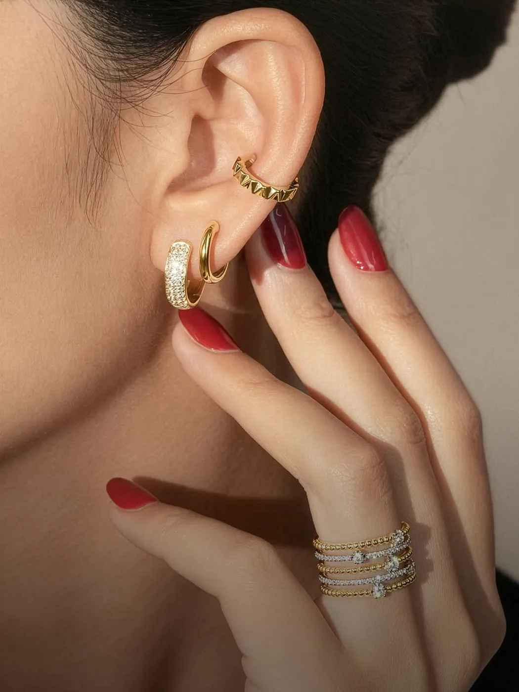 Close-up of ear with gold earrings and hand with red nails and stacked rings. Explore earrings at McGee & Company.