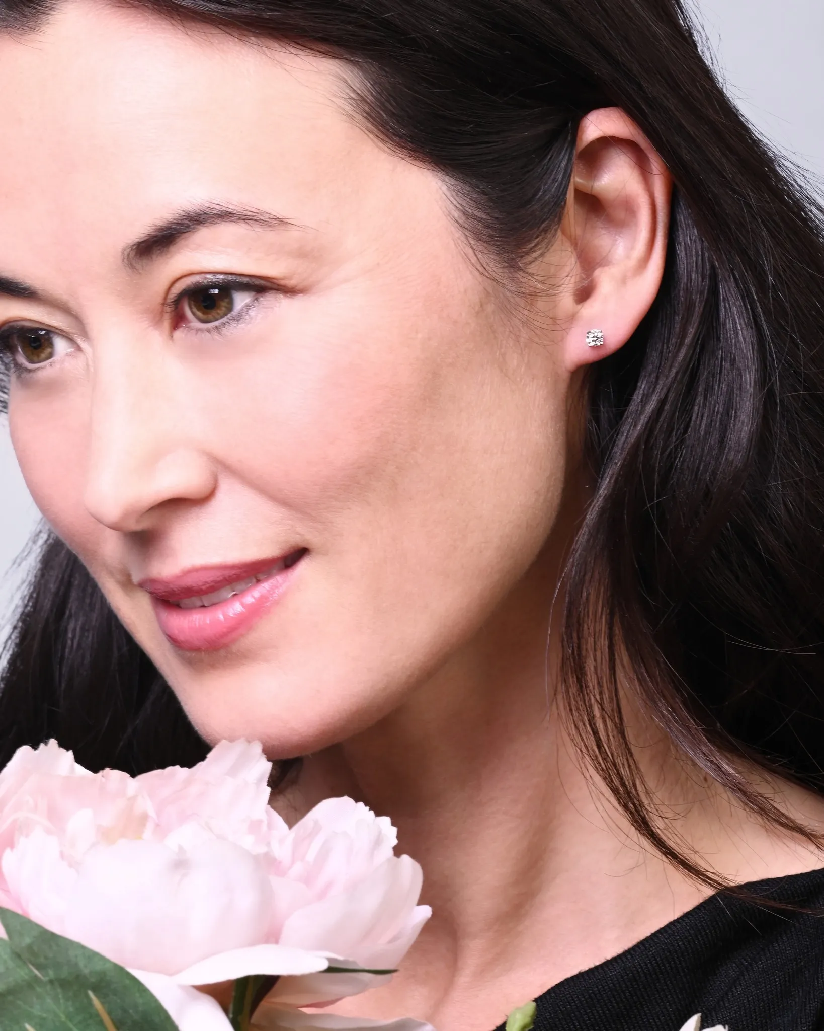 Woman with diamond stud earrings holding a pink flower, promoting diamond studs. Click to shop all diamond studs.