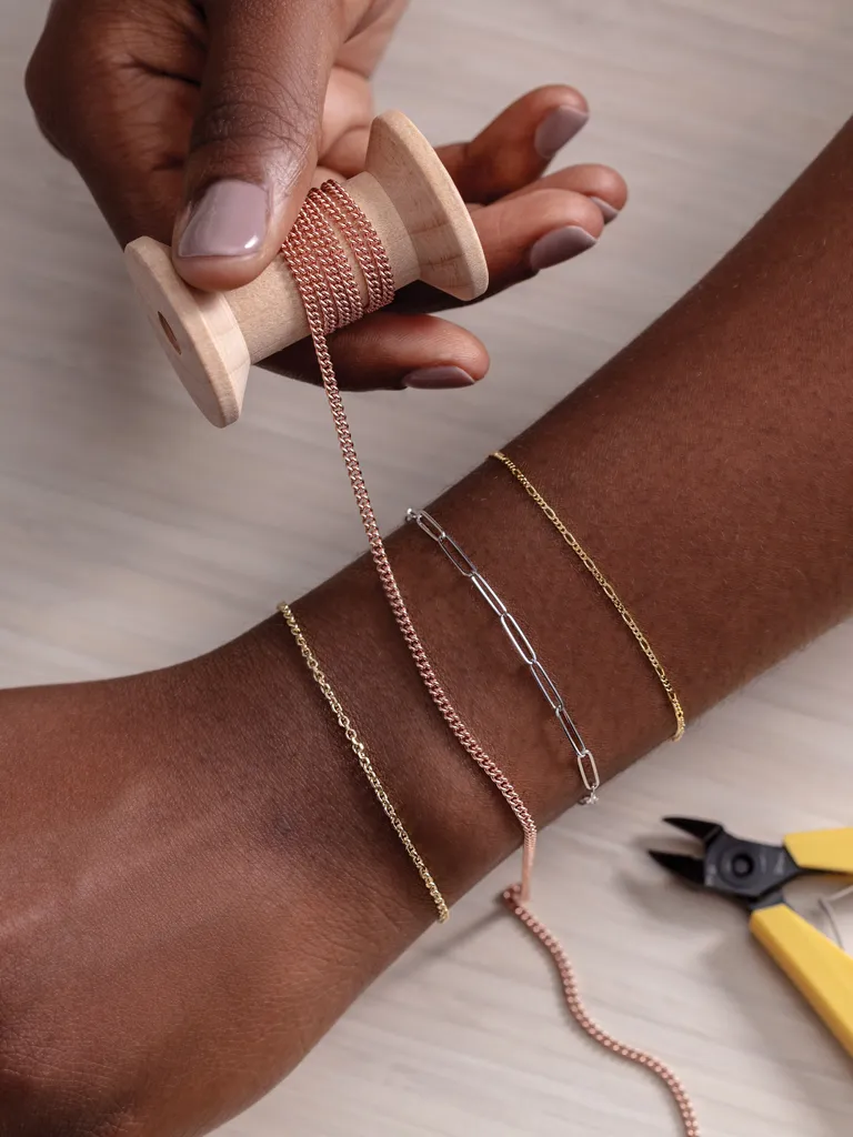 Hand holding a spool of chain while crafting jewelry, showcasing gold and silver bracelets on a wrist.