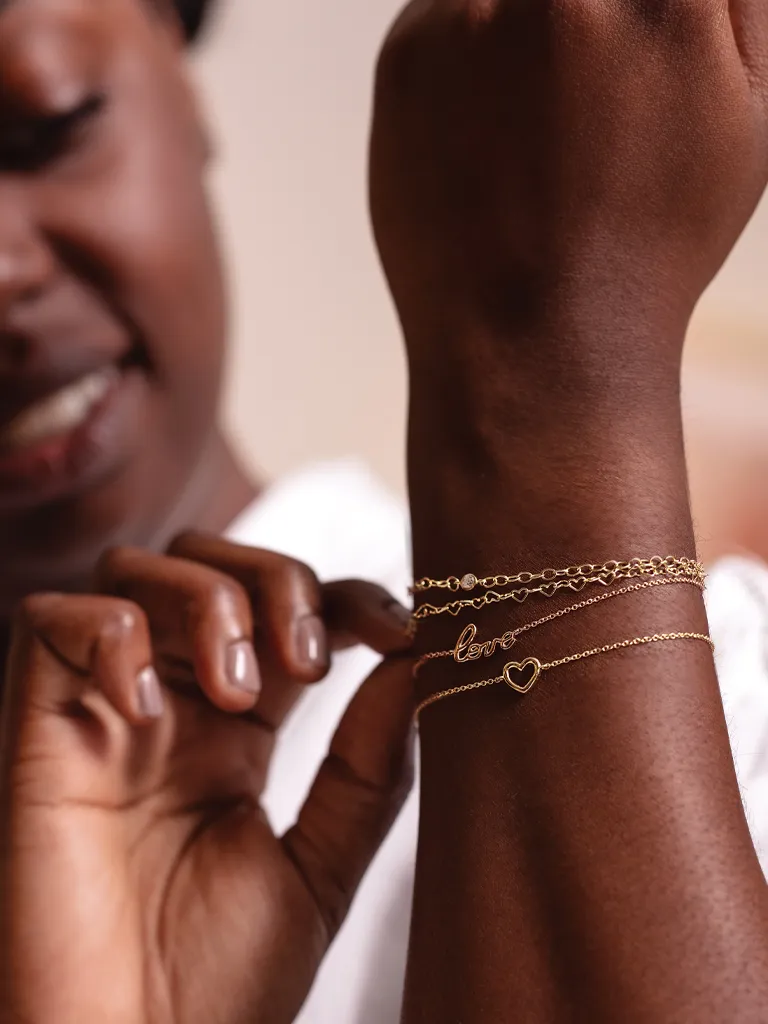A woman showcases three gold bracelets on her wrist, featuring a heart and the word 