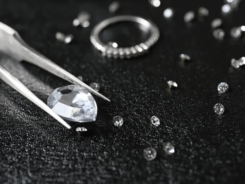 A pear-shaped diamond held by tweezers, surrounded by smaller diamonds on a dark surface.