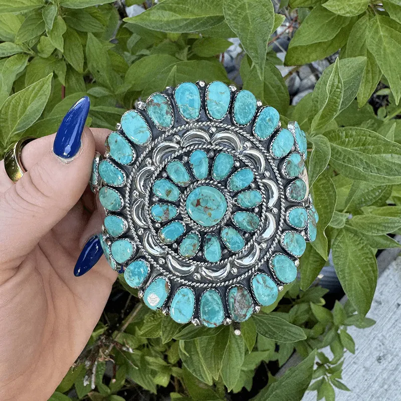 Turquoise and silver decorative piece held in hand, surrounded by green leaves.