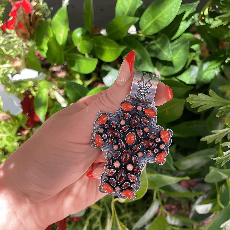 Hand holding a silver cross pendant with red stones, surrounded by greenery.