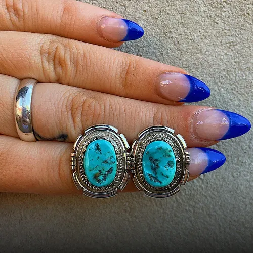 Turquoise earrings set in silver, displayed on a hand with blue-tipped nails. 