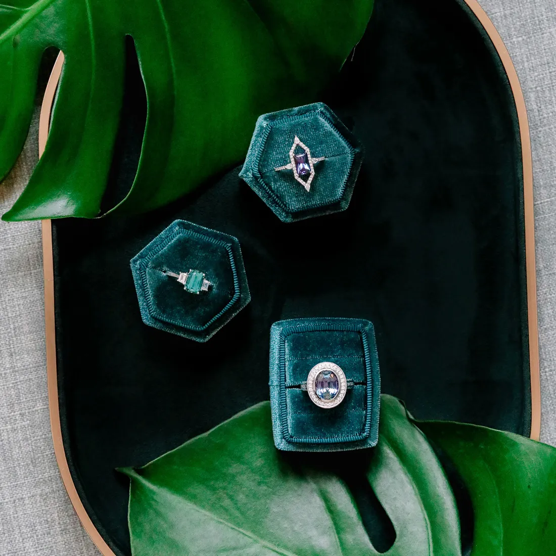 Three elegant gemstone rings displayed in green velvet boxes on a dark tray with tropical leaves.