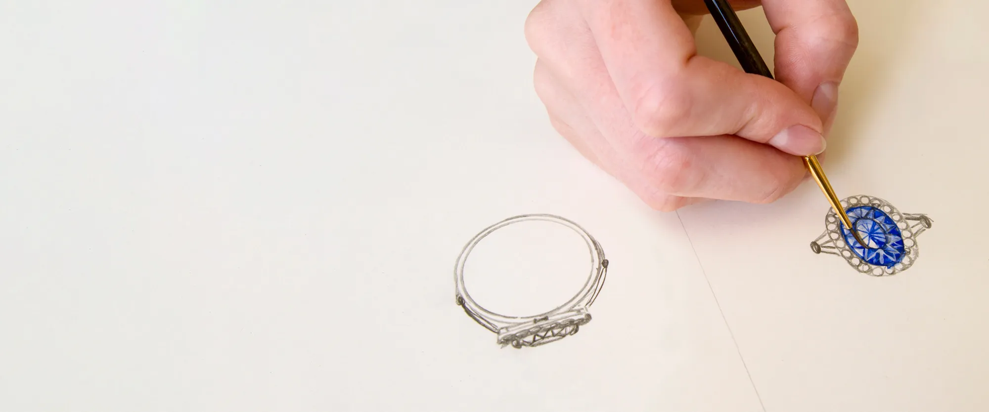 A hand painting a blue gemstone ring design on paper, showcasing custom jewelry creation at Michael & Son's Jewelers.