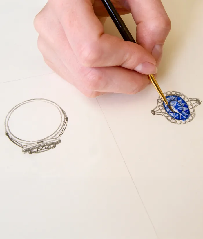 A hand painting a blue gemstone ring design on paper, showcasing custom jewelry creation at Michael & Son's Jewelers.