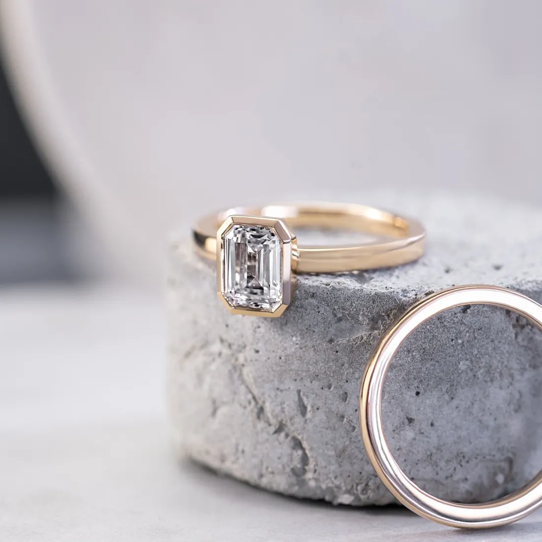 Elegant gold engagement ring with an emerald-cut diamond, paired with a simple gold band, displayed on a concrete pedestal.