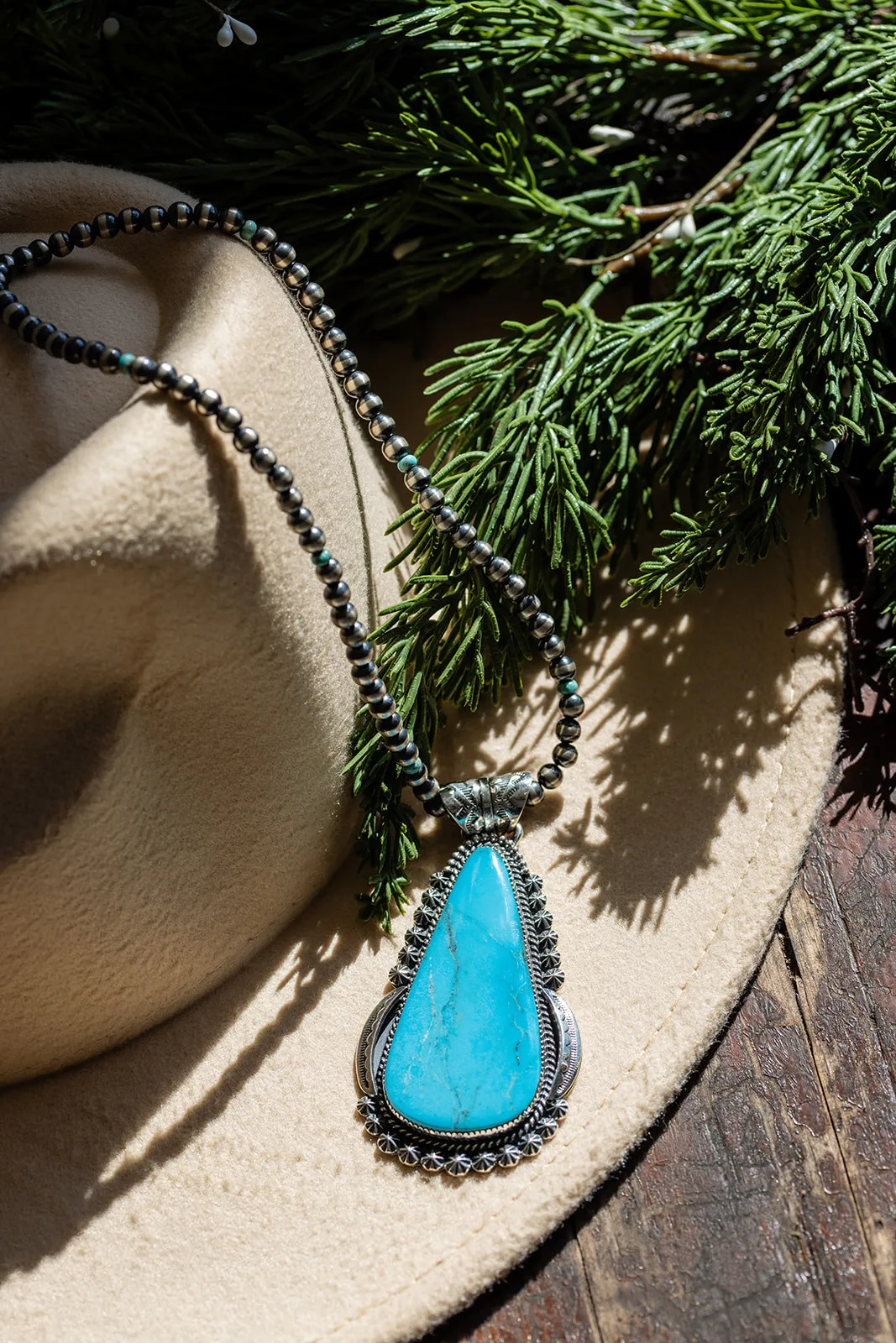 A turquoise pendant on a silver bead necklace resting on a cream hat with pine boughs