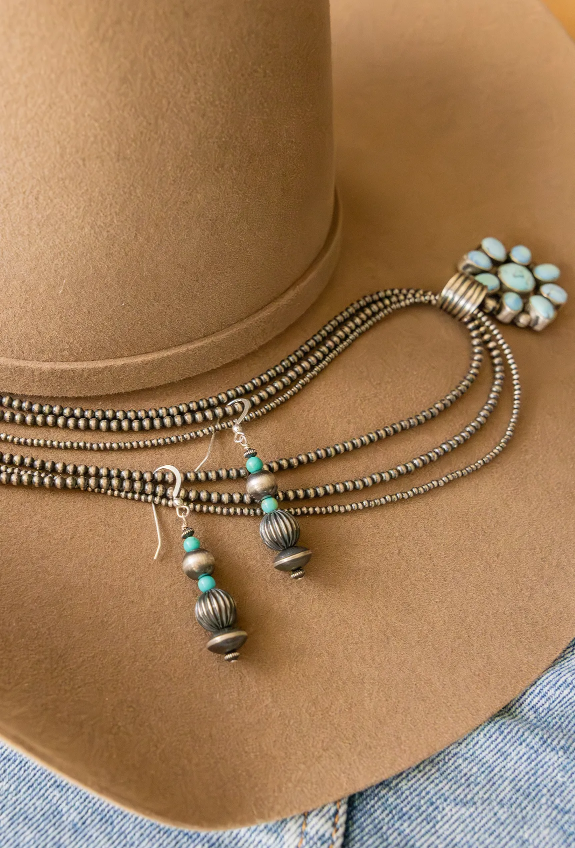 A tan cowboy hat adorned with layered silver and turquoise jewelry, including a necklace and earrings.