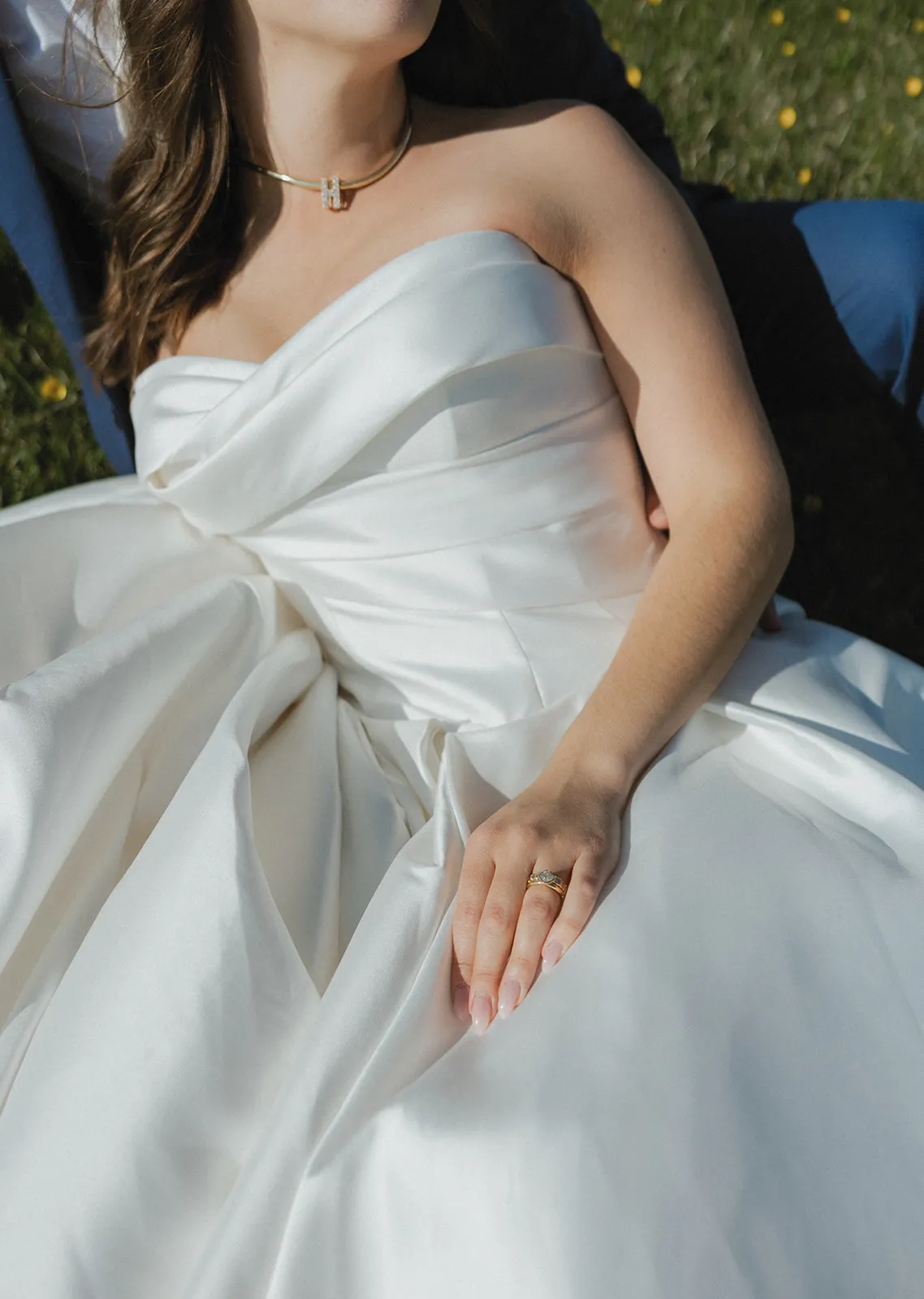 Bride in a white satin gown with a gold necklace and ring, lying on grass.
