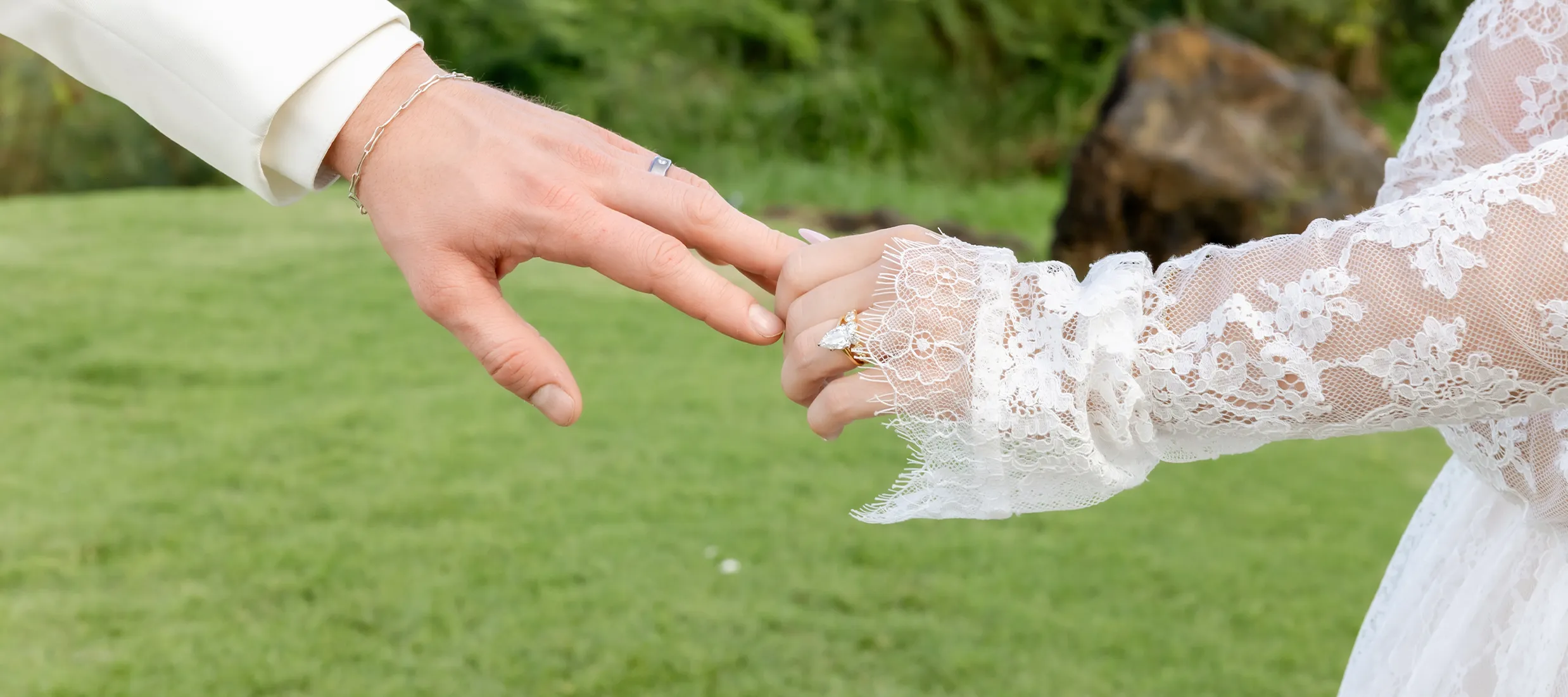 A bride and groom's hands touching, with the bride wearing a lace sleeve and a ring, set against a grassy background.