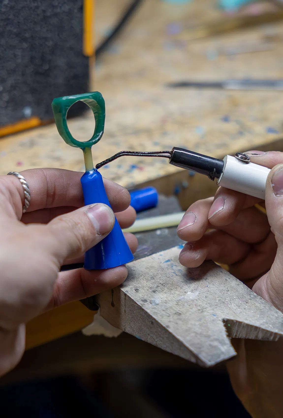 A person using a soldering tool on a green wax ring model at a jeweler's workbench.
