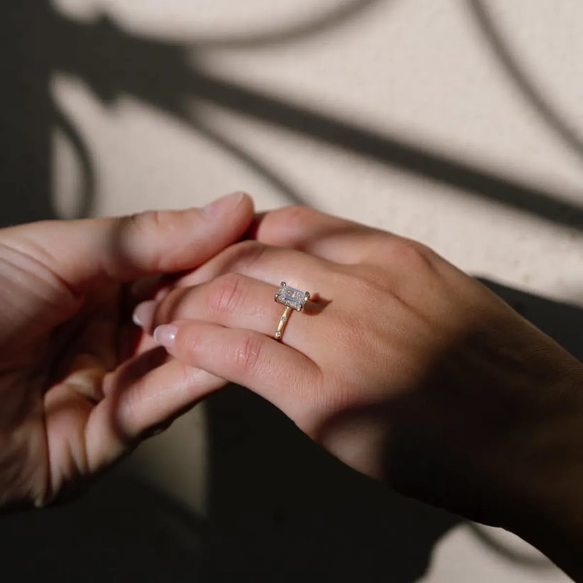 Two hands holding each other, one wearing a rectangular diamond ring, with shadows on a light background.