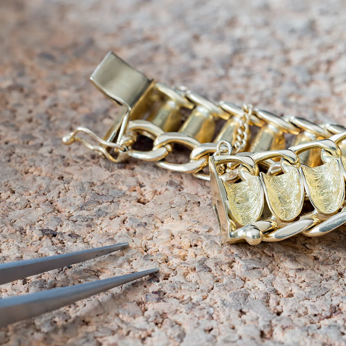 Gold bracelet with intricate design on cork surface, tweezers nearby.