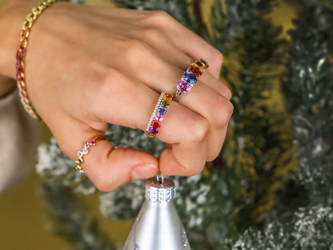 Two hands adorned with colorful rings and bracelets, holding Christmas ornaments against a festive backdrop.