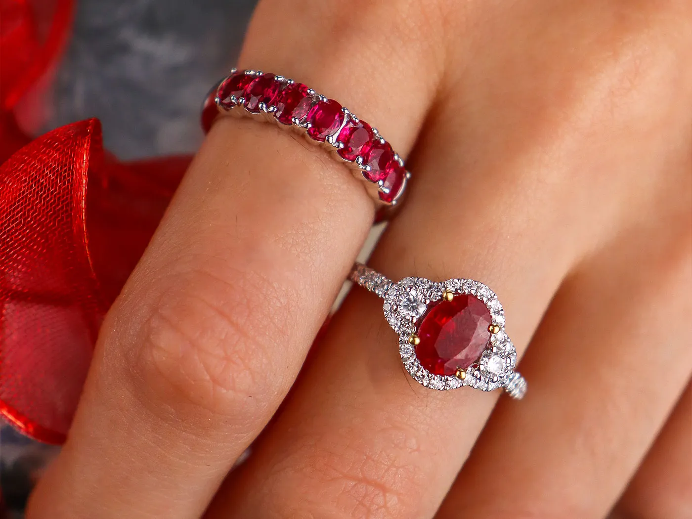 Two elegant rings featuring rubies and diamonds on a hand, with a red ribbon in the background. Click to shop rings.