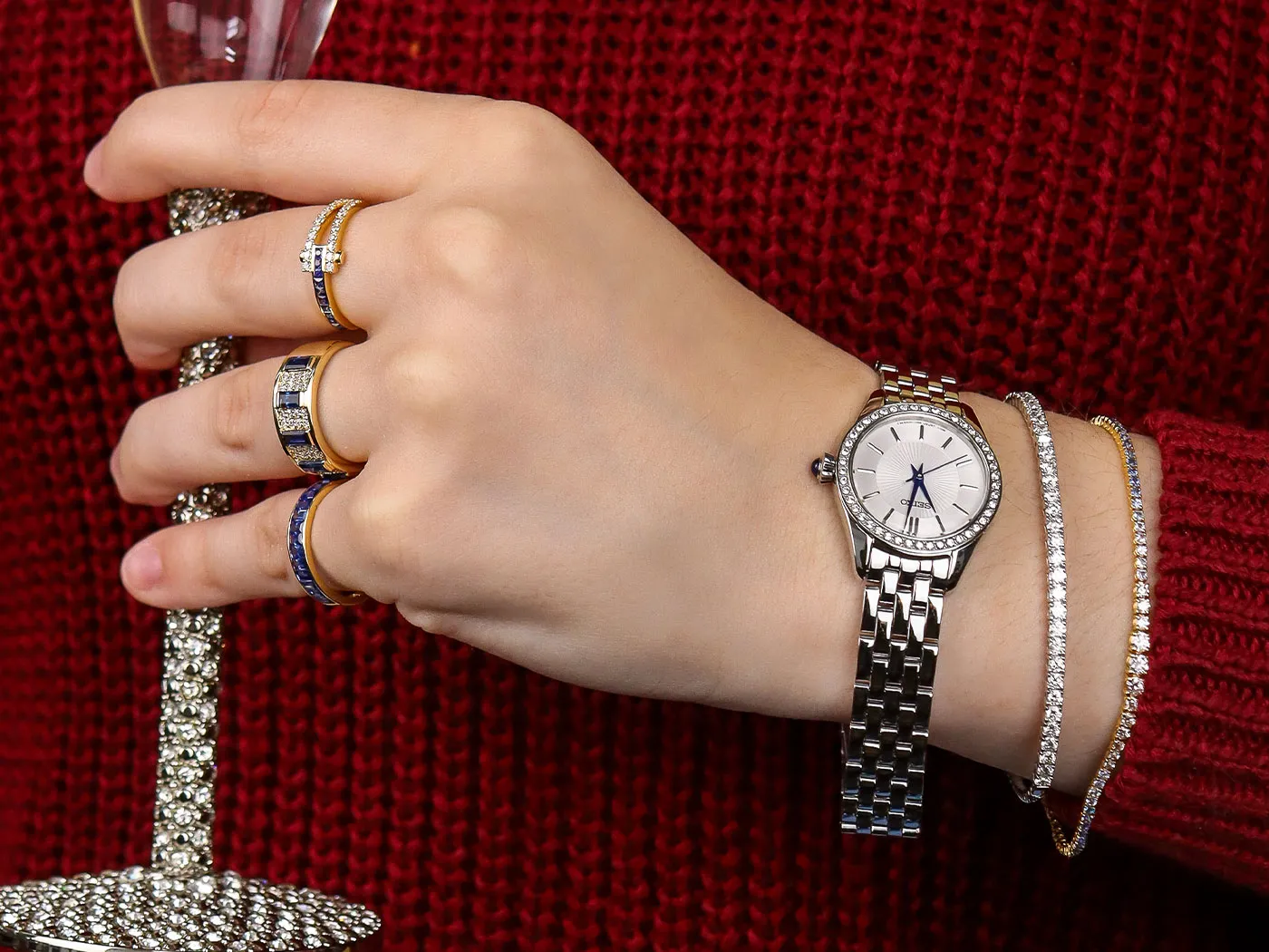 Hand adorned with a silver watch and multiple rings, holding a sparkling glass, against a red sweater backdrop. Shop all jewelry