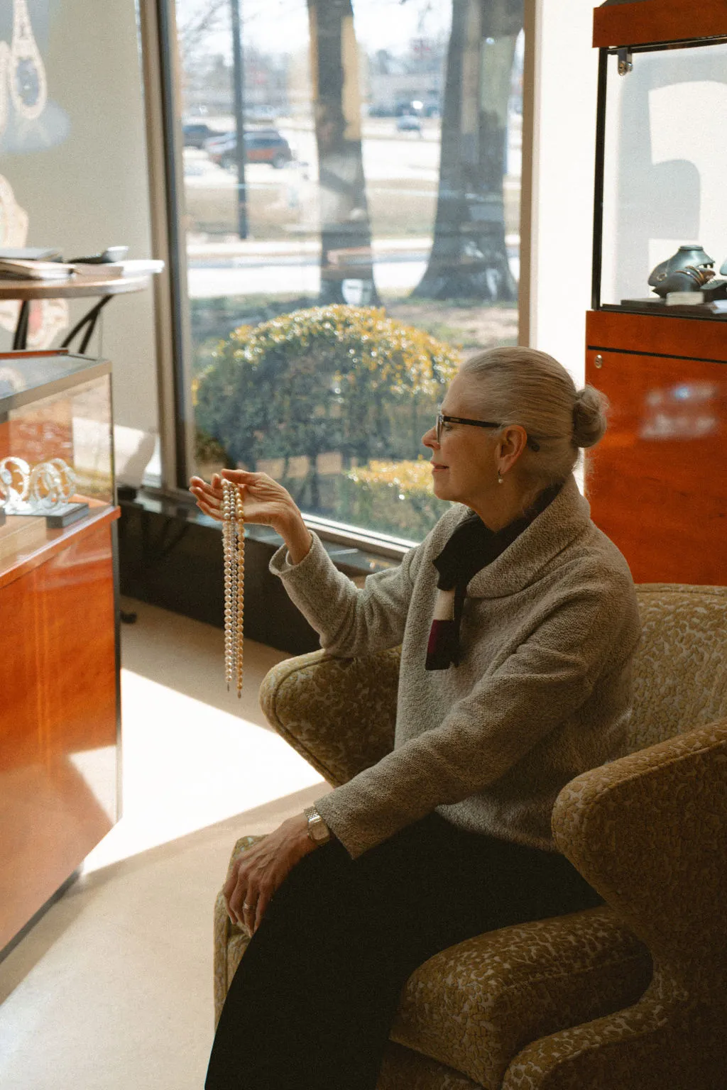 A woman in a jewelry store admires a pearl necklace by a window.