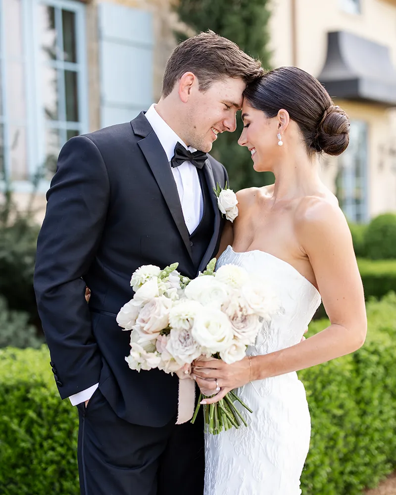 Bride and groom smiling, holding a bouquet, in a garden setting.