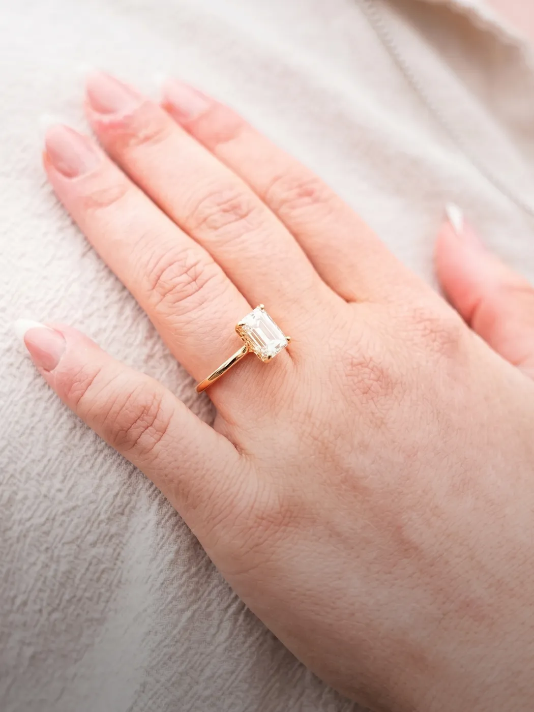 A hand wearing a gold ring with a rectangular diamond on a light fabric background.