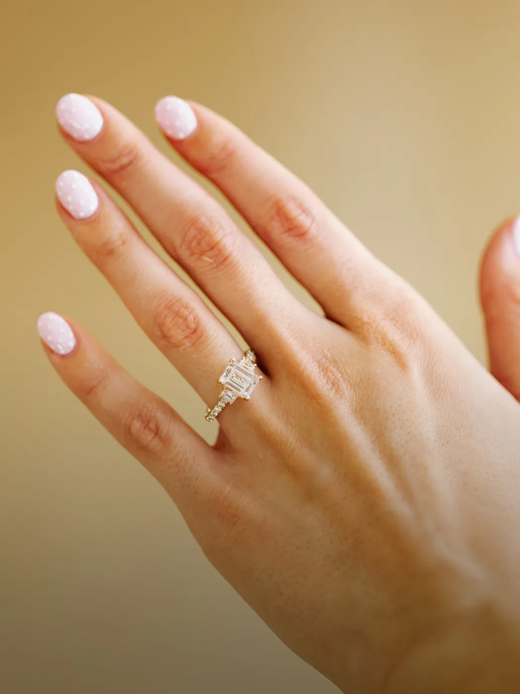 Hand with a diamond engagement ring and polka dot nails against a beige background.