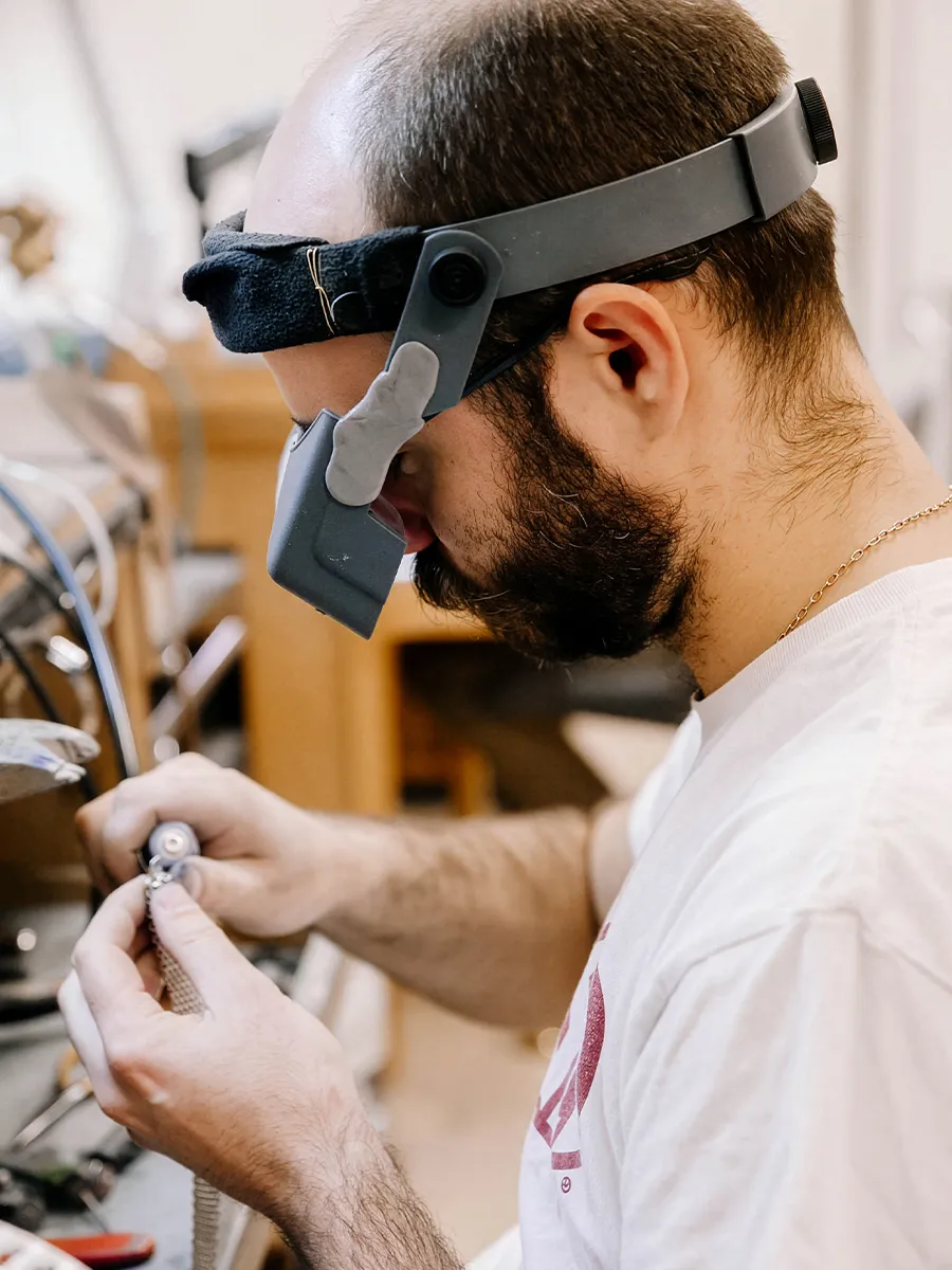 A jeweler wearing magnifying glasses works on a piece of jewelry at a workbench.