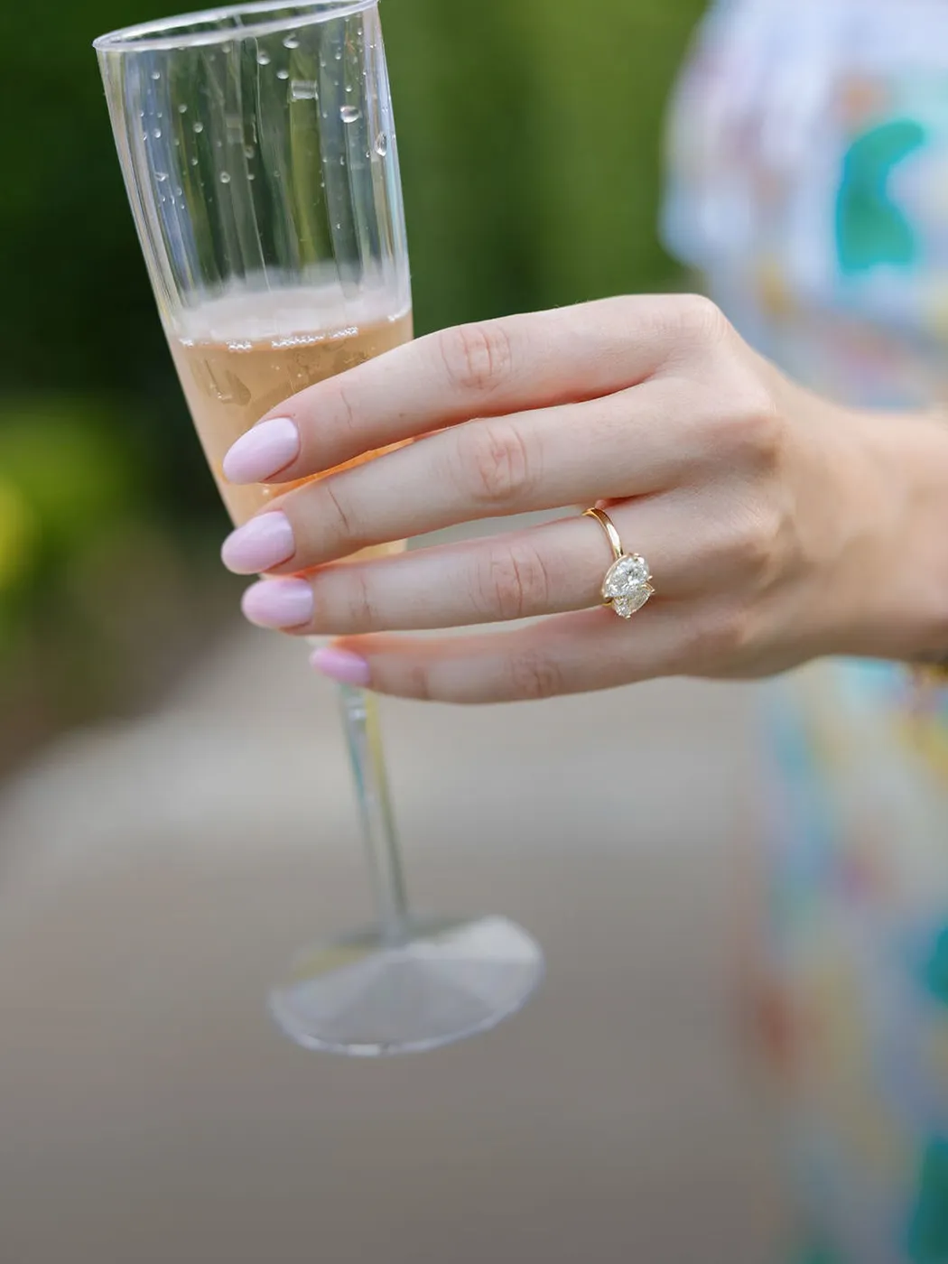 A hand with a diamond ring holds a champagne flute outdoors.