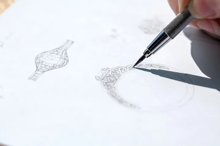 A hand sketches intricate jewelry designs on paper, showcasing creativity and craftsmanship at Monarch Jewelry in Winter Park.