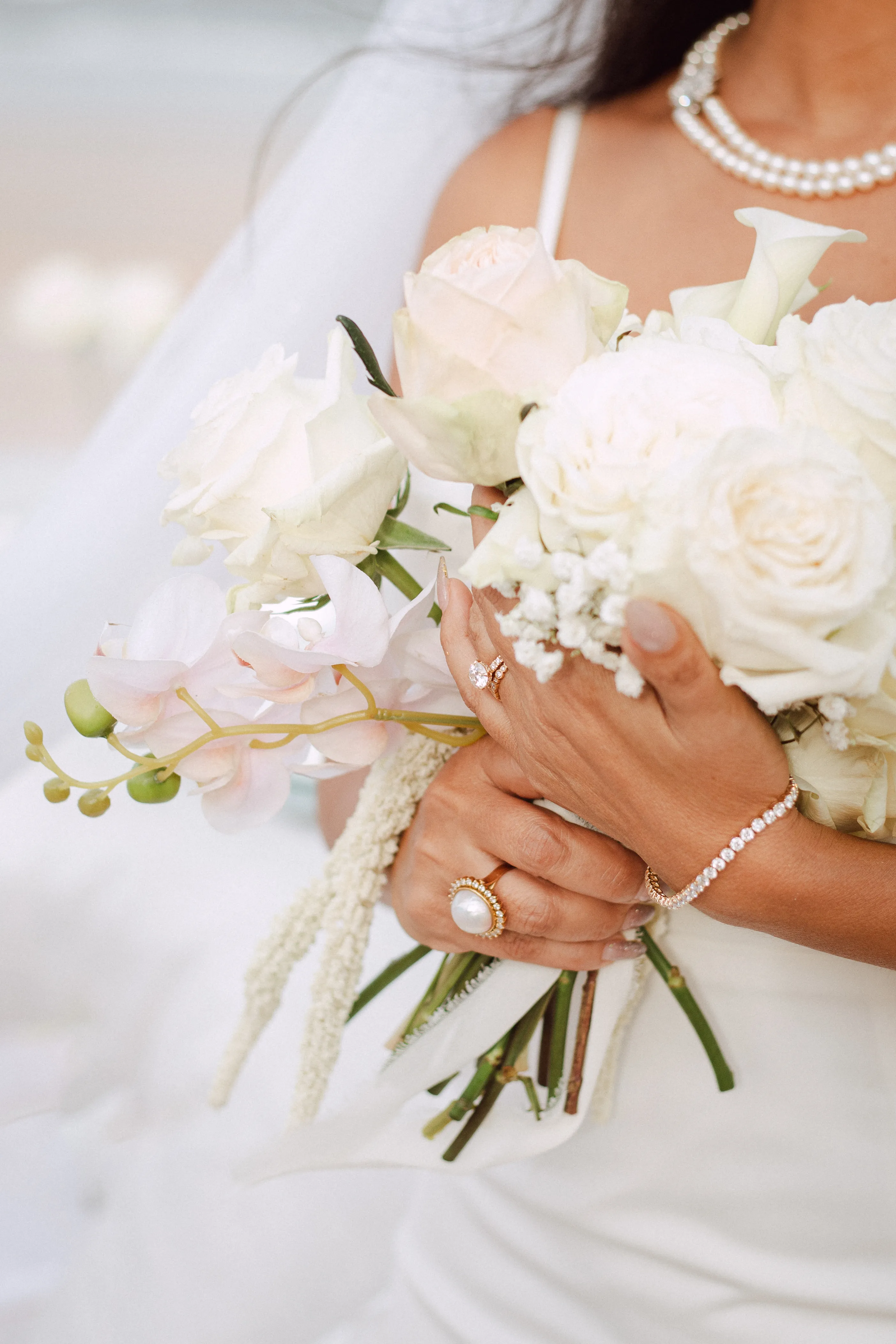 A bride holding a bouquet of white roses and orchids, adorned with elegant jewelry. Visit Monarch Jewelry for more.