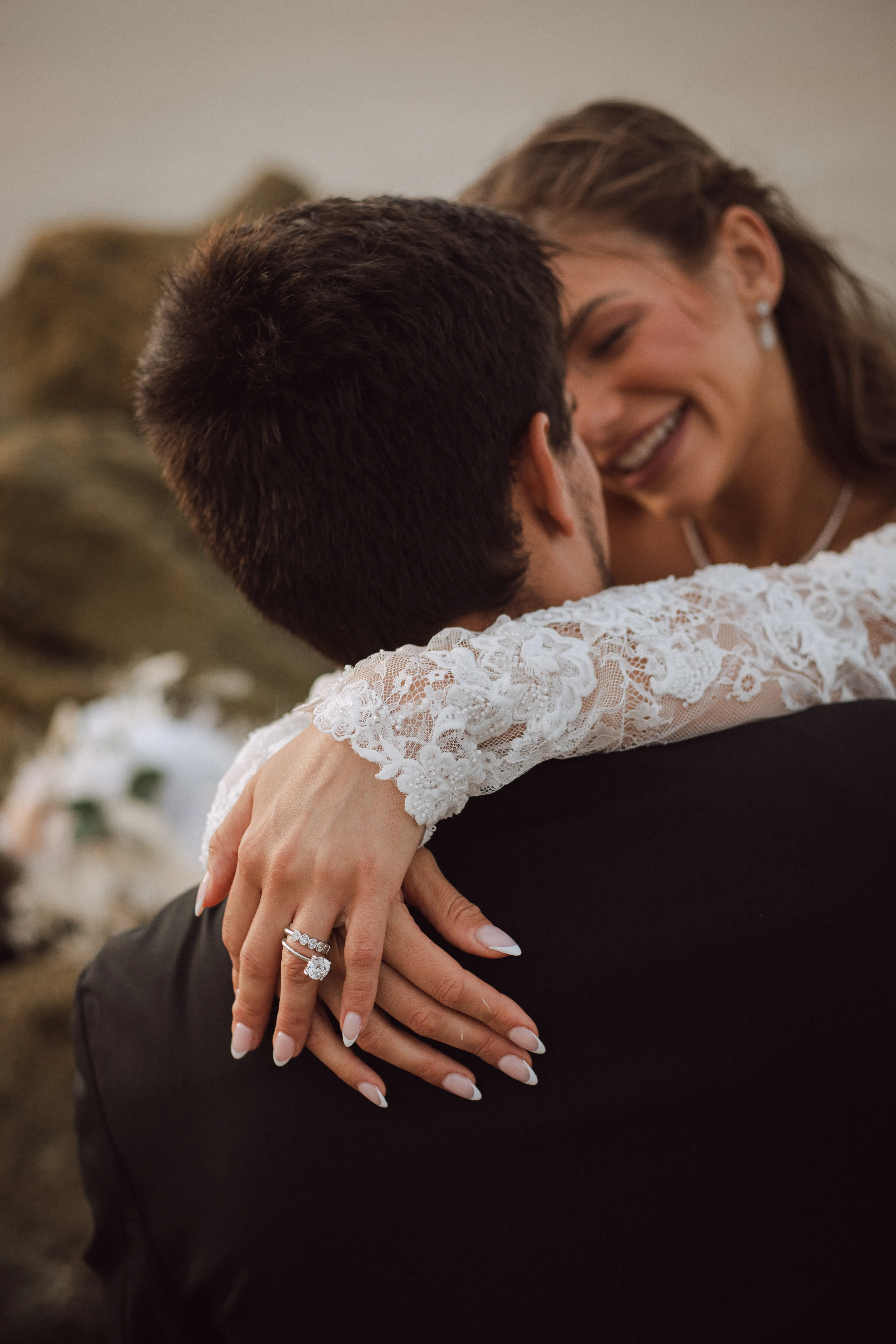 A bride embraces a groom, showcasing a lace sleeve and diamond ring.