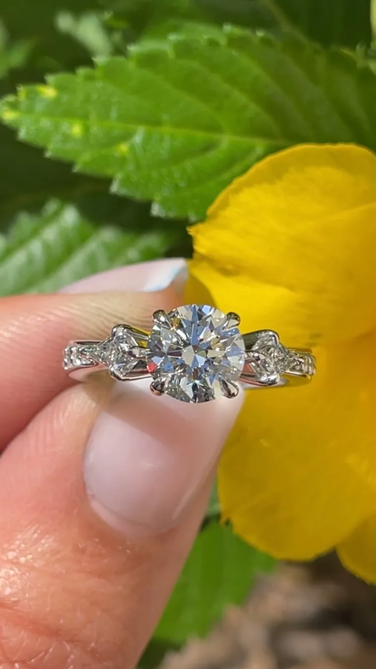 A hand holding a sparkling engagement ring with a round diamond, set against green leaves and a yellow flower background.
