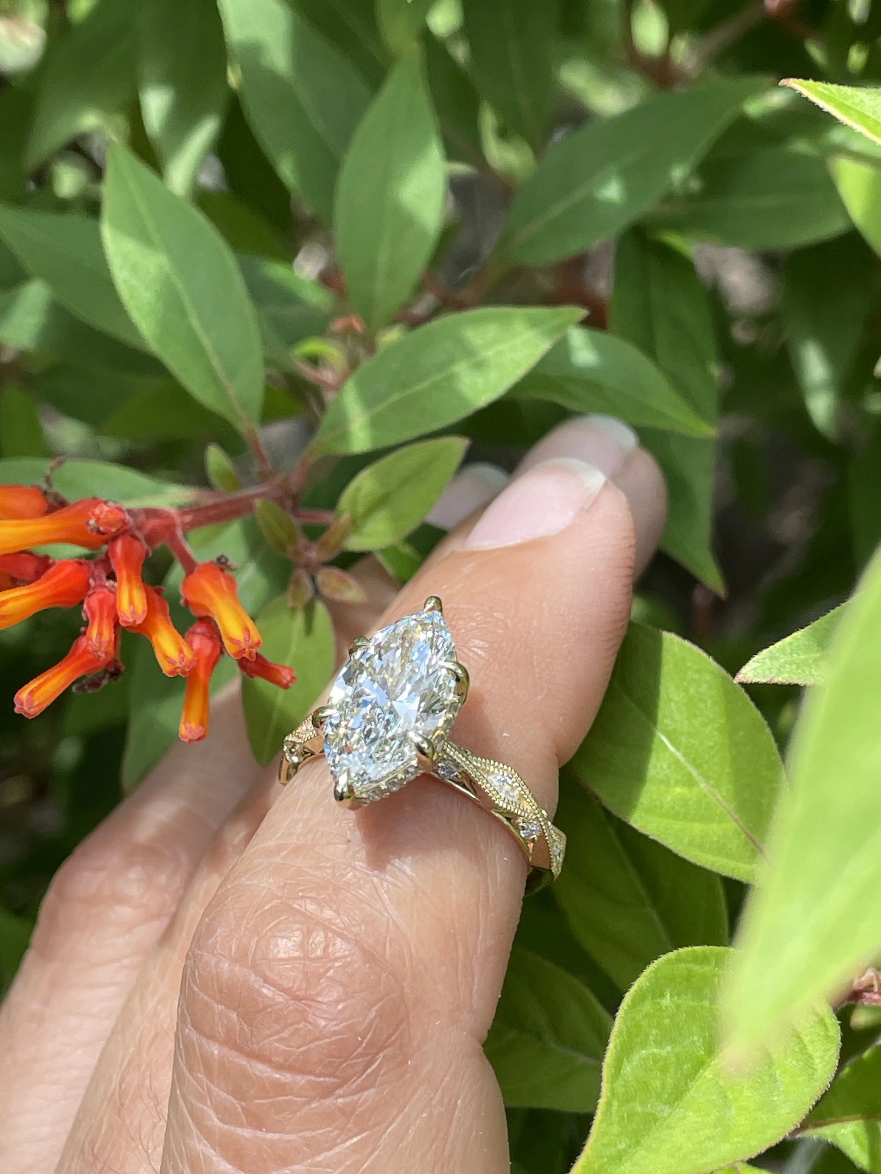 A hand holding a marquise diamond engagement ring among green leaves and orange flowers. Visit Monarch Jewelry in Winter Park.