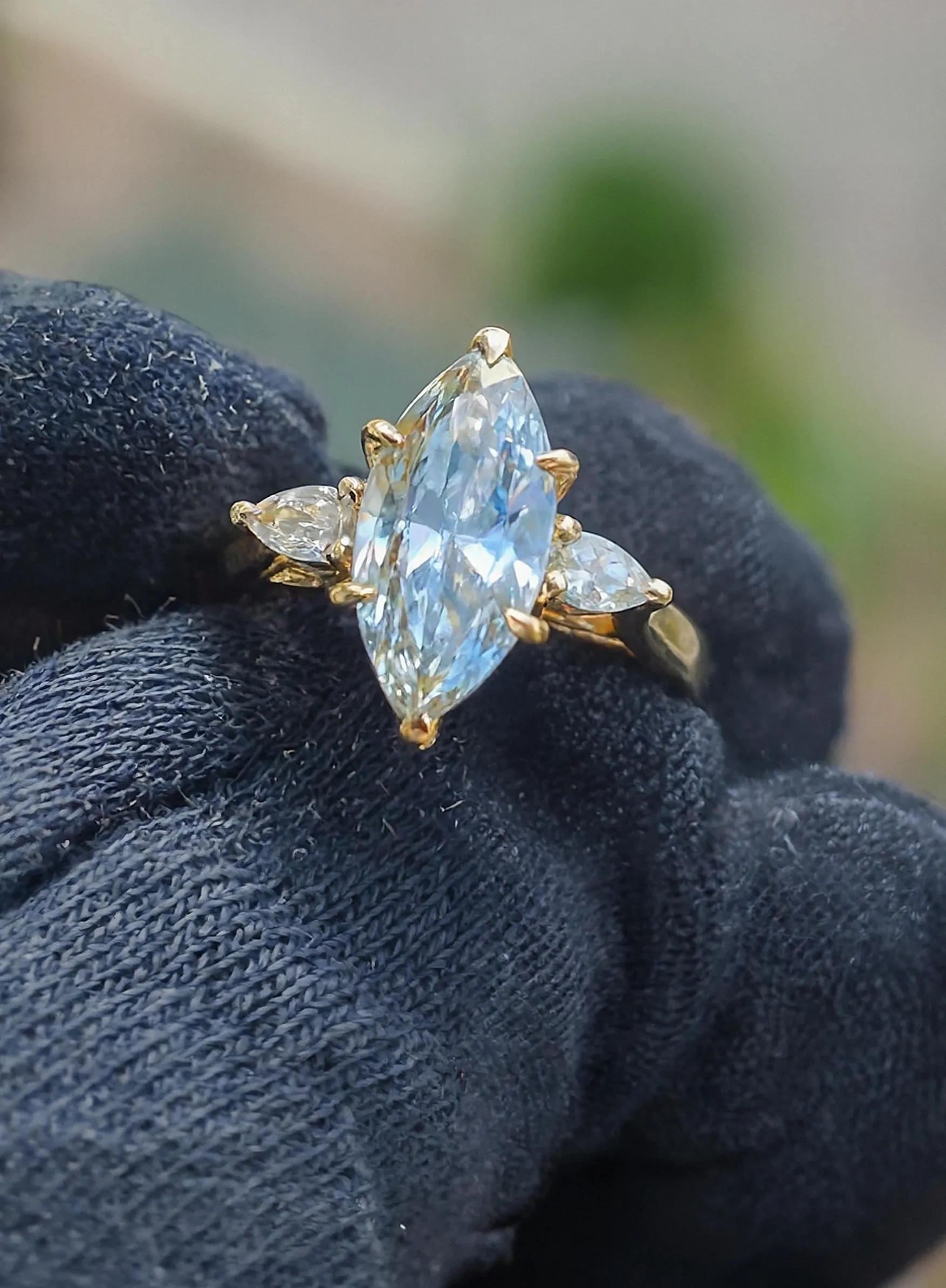 A stunning marquise-cut blue gemstone ring with two smaller side stones, set in gold, held by a gloved hand.