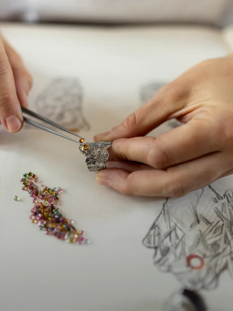 A person using tweezers to place a gemstone on a jewelry design sketch, surrounded by colorful beads.