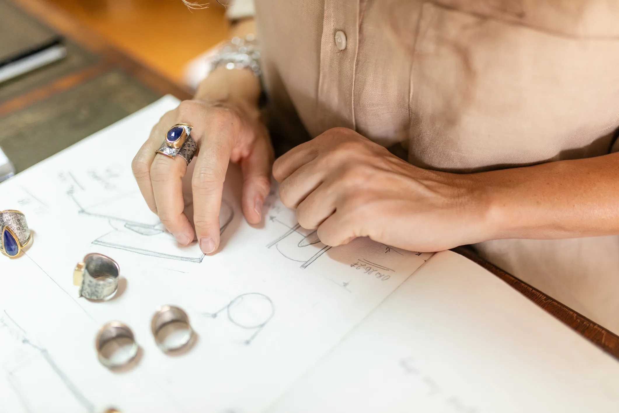 Hand sketching a colorful necklace design with a heart, surrounded by other jewelry sketches on a workspace.