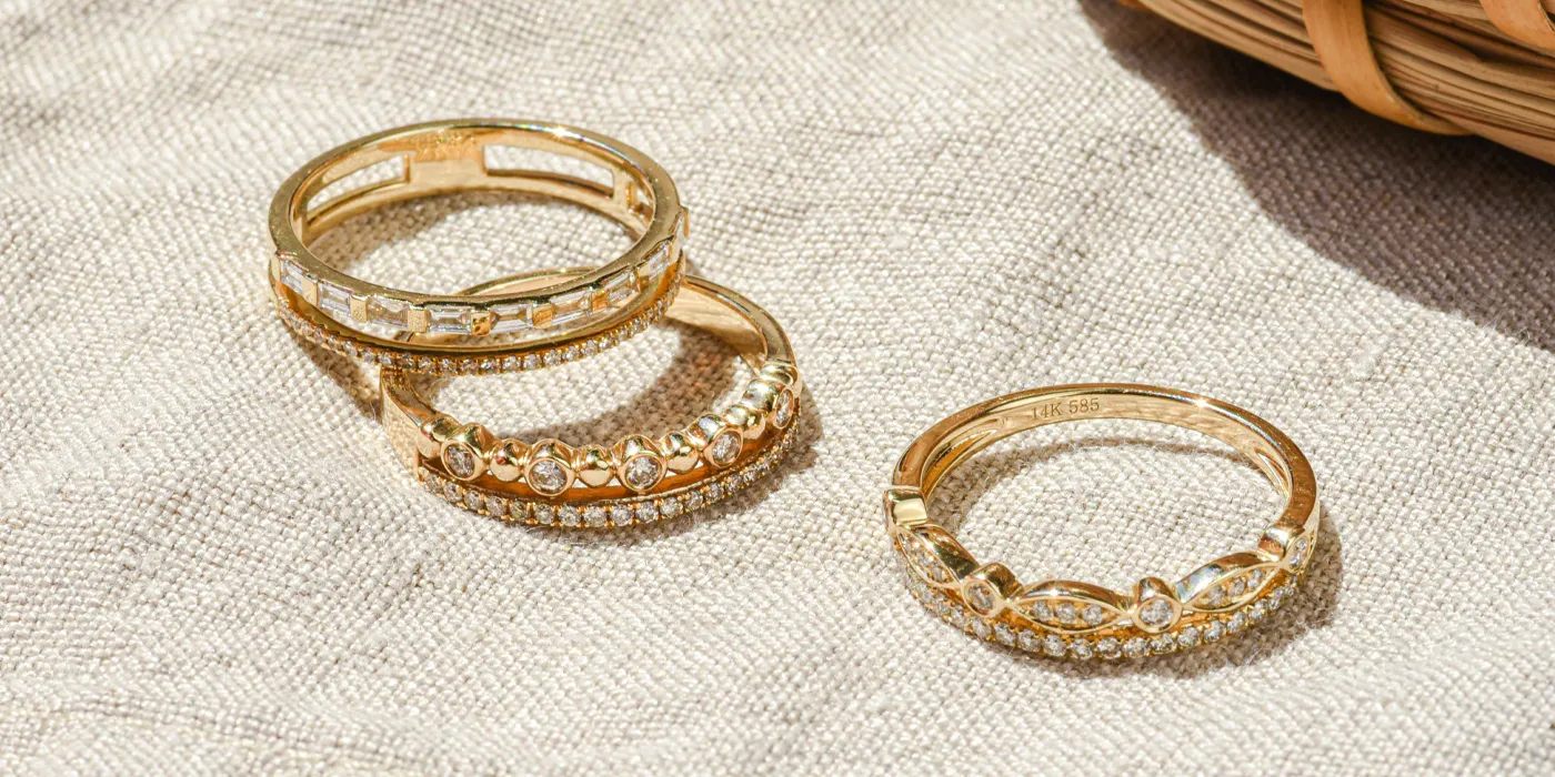 Three elegant gold rings with sparkling stones on a textured fabric background. Visit Monarch Jewelry in Winter Park.