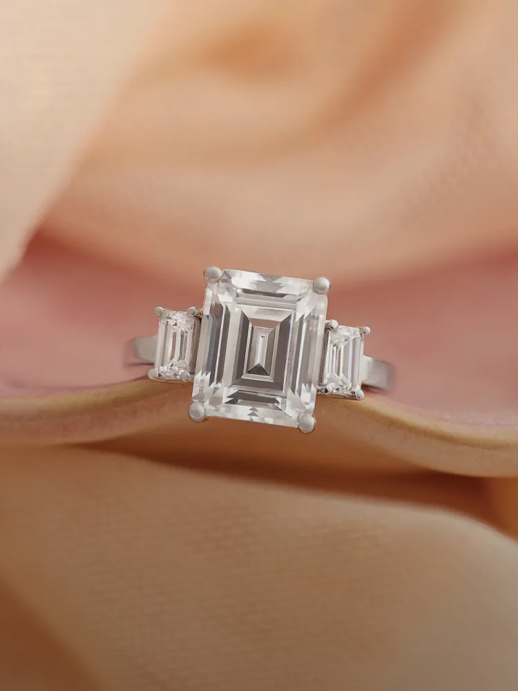 Elegant engagement ring featuring a central emerald-cut diamond flanked by two smaller diamonds. Shop at Monarch Jewelry.