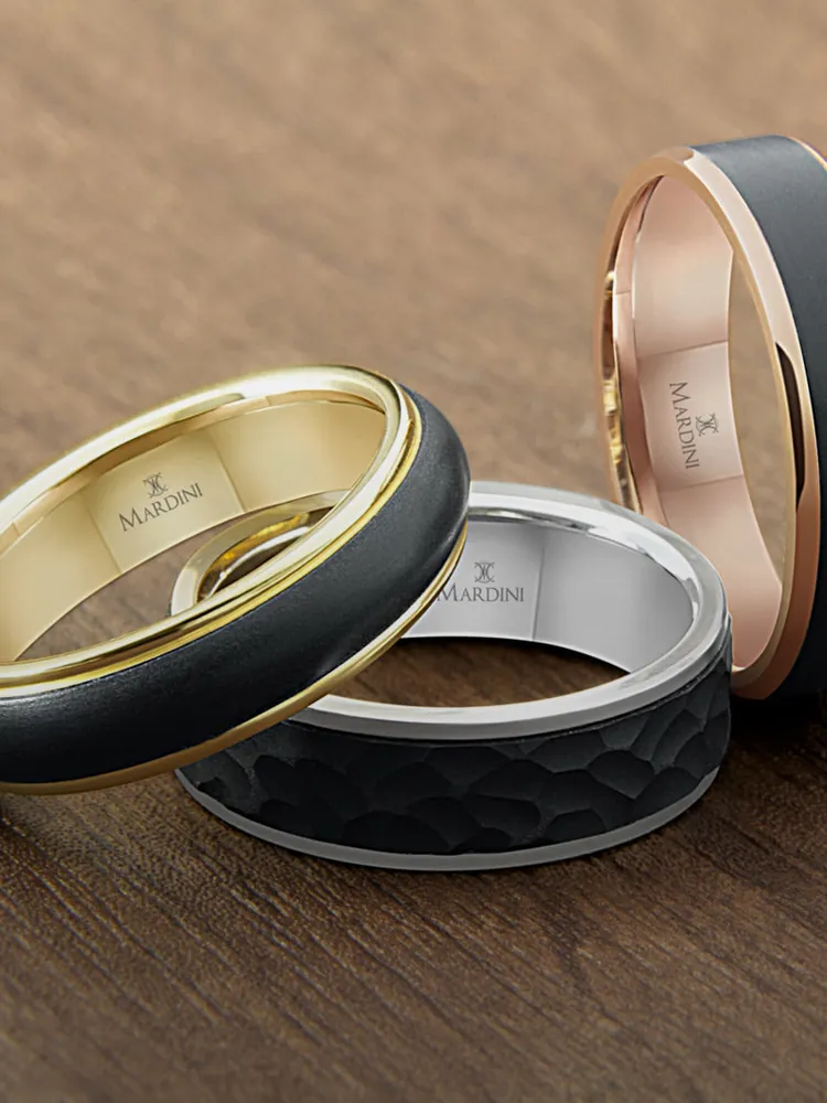 Three stylish men's wedding bands in gold, silver, and black, displayed on a wooden surface. Shop Men's Wedding Bands.